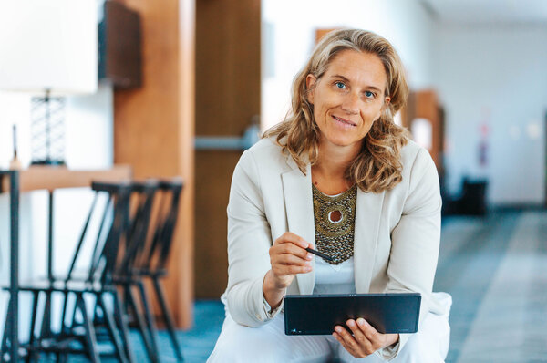 Corina Hoch sitzt in einem modernen, hellen Innenraum und lächelt. Sie hält ein Tablet in der Hand und hat einen Stift bereit. Ihr Outfit ist elegant und professionell, passend zu einem Arbeits- oder Beratungssituation. Im Hintergrund sind Möbel und eine helle Wand zu erkennen.