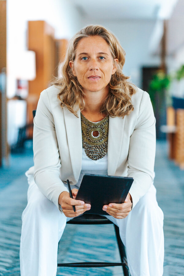 Corina Hoch sitzt auf einem Stuhl und hält ein digitales Gerät in der Hand. Sie trägt einen hellen Blazer und einen auffälligen Halsreifen. Ihr Blick ist fokussiert und direkt. Im Hintergrund sind unscharfe Elemente eines modernen Innenraums zu erkennen.